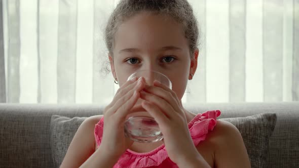 Cute Baby Girl Drinking a Glass of Water Sitting on the Couch at Home