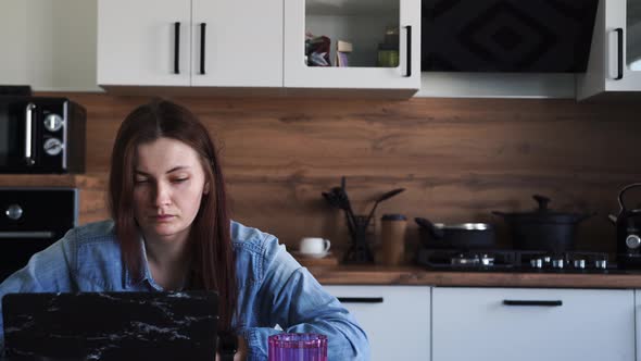 Woman Working at Laptop While Sitting at the Table in the Kitchen at Home