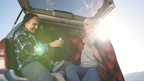 Adventurous Young Women Sitting in the Car Trunk, Holding Thermos and Drinking Coffee