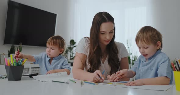 A Young Mother with Two Children Sitting at a White Table Draws Colored Pencils on Paper