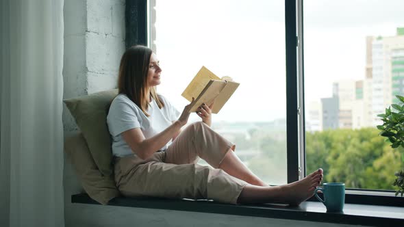 Beautiful Girl Reading Book Turning Pages Sitting on Window-sill at Home