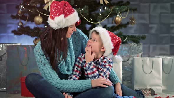 Smiling Young Mother and Little Cute Child Hugging Sitting Together and Talking Near Christmas Tree