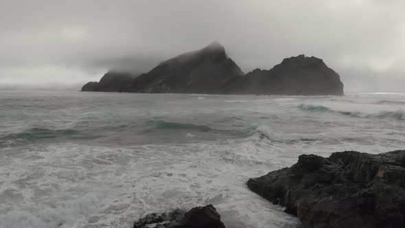 Aerial forward over sea with big rock shrouded in mist on background, Ponta da Calheta