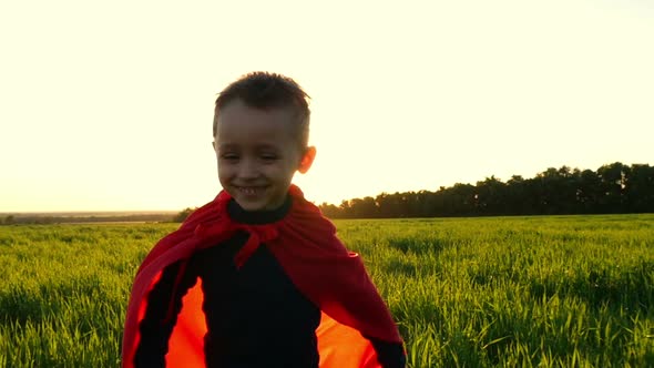 A Happy Child in a Superhero Costume in a Red Cloak Runs on Green Grass on a Sunset Background in