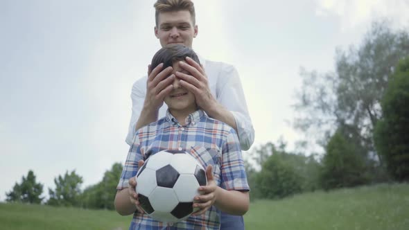 Portrait of the Cute Boy Looking in the Camera, His Father or Older Brother Covering His Eyes From