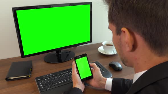 An office worker sits at a desk and looks at a computer and a smartphone with green screens