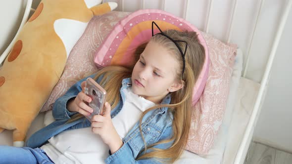 a Pretty Little Girl in a Rim with Cat Ears with a Phone in Bed