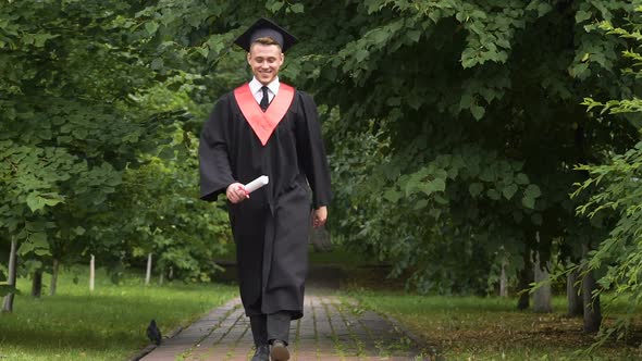 Smiling Young Man in Academic Dress Carrying Diploma in Hands, Jumping With Joy