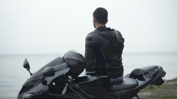 Back View of Confident Middle Eastern Male Biker Standing with Motorcycle on River Bank and Thinking