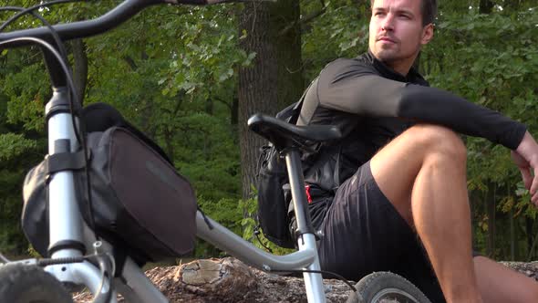 A Young Handsome Cyclist Sits on a Log Next To His Bike in a Forest and Looks Around