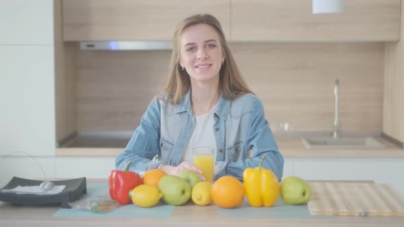 A Beautiful Young Woman with Glass of Juice is Sitting at Kitchen Table