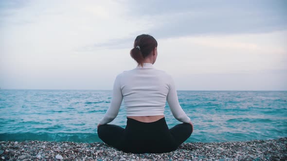 A Woman Sits on the Pebble Beach in the Lotus Position and Looks at the Sea