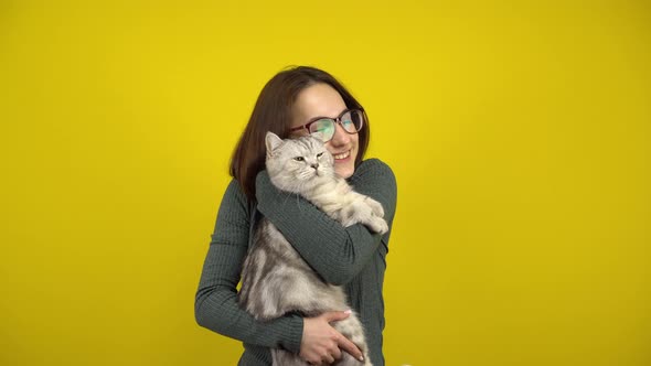 A Young Woman Holds a Cat in Her Arms and Hugs Him on a Yellow Background. Woman with Glasses and a