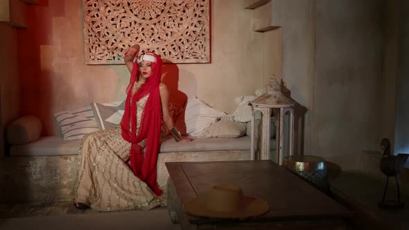 a Woman in a Turban a Red Scarf and a Sequined Dress is Sitting on a Sofa with Pillows in the Sunset