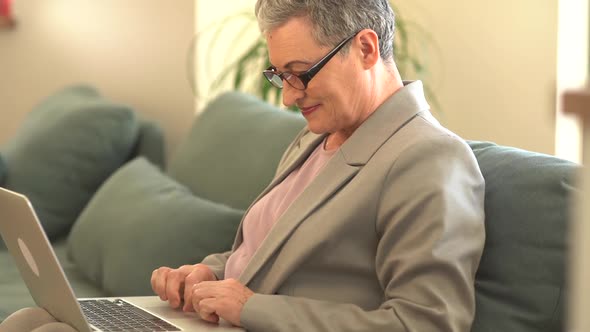 Beautiful Middleaged Business Lady Wearing Glasses and a Gray Business Suit is Working with a Laptop