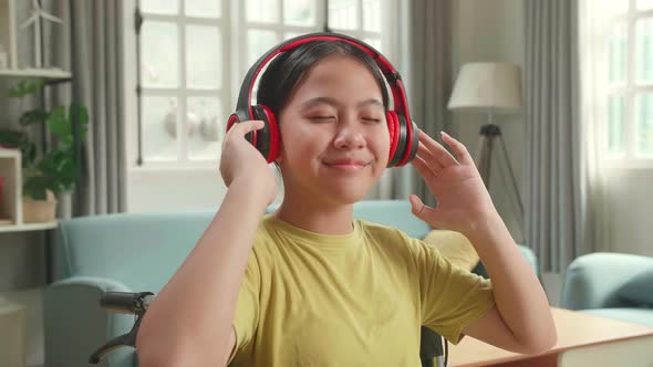 Asian Kid Girl Sitting In A Wheelchair Listening To Music With Headphones And Dancing In Living Room