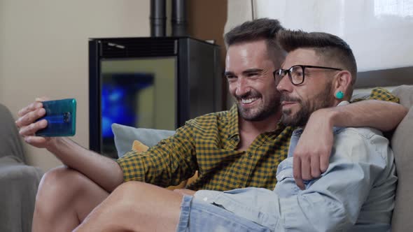 Gay Couple Sitting in the Sofa At Home Making Video Call On Mobile Phone