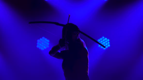 Silhouette of Dancing Joan of Arc with Bow and Arrow on Stage in a Dark Studio with Smoke and Neon