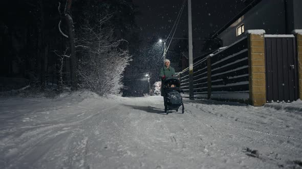 Young Mother Pushing Baby Stroller and Walking at Winter Park During Snowfall