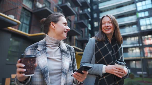 Two Happy Women Walking with Takeaway Coffee and Talking with Interest Among Themselves in the