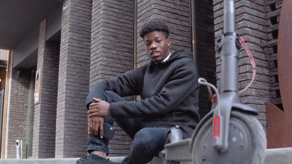 A Young African American Man in a Black Sweater and Jeans and Sneakers Sits on the Sidewalk Near a