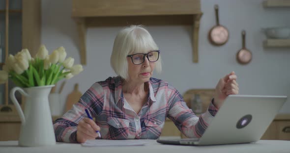 Senior Teacher in Glasses Writing with Pen on Papers Looking at Laptop. Online Education and