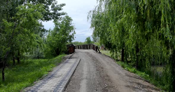 Dirt Road With Willow Trees Towards The Wooden Bridge