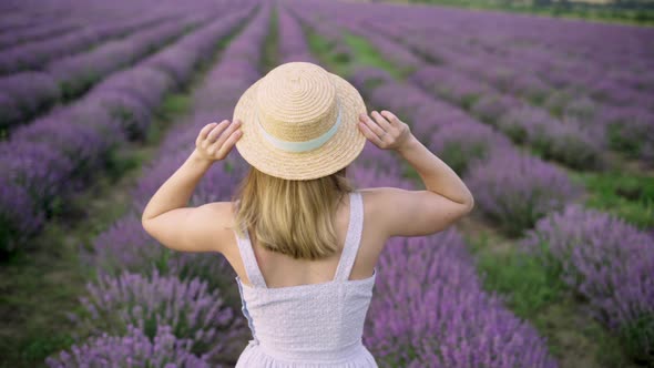 Rear View of a Young Happy Woman Holding a Straw Hat and Walking at Sunset or Dawn on a Lavender