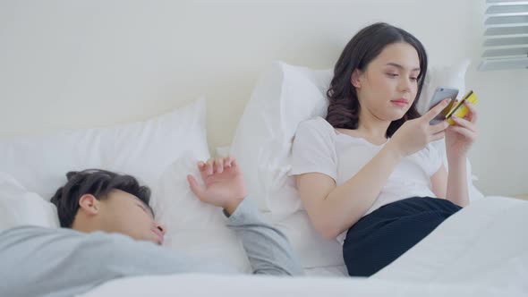 Asian woman holding credit card enjoy shopping online on bed while her husband sleeping.