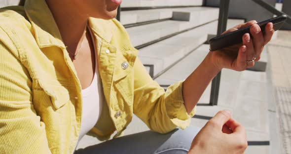 Happy caucasian woman sitting on stairs, using smartphone on sunny day