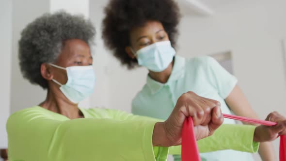 African american female physiotherapist wearing face mask helping senior female patient exercise