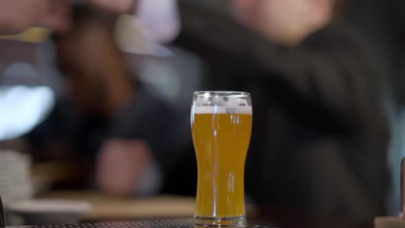 Closeup Pale Lager in Beer Glass on Bar Counter with Blurred Men Receiving Order Drinking at