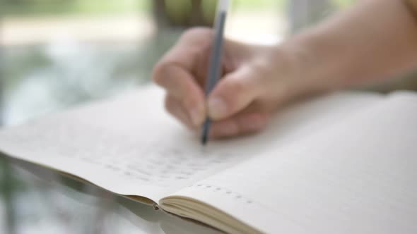 Woman Writes in Notebook with Pen Sitting at Glass Table