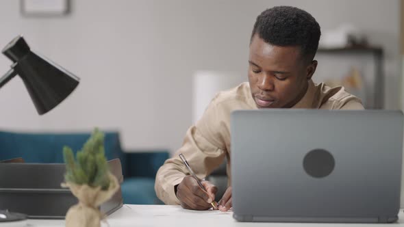 Afroamerican Student is Working in Apartment Writing on Paper Sitting at Table with Laptop Online