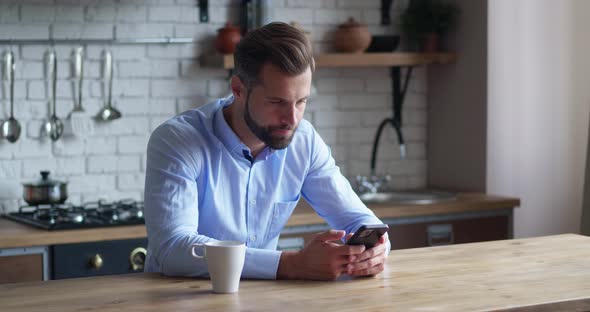 Bearded Male Millennial Professional Holding Modern Smartphone Texting Message in Kitchen