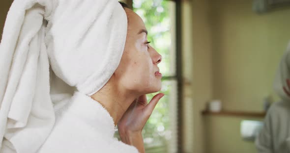 Biracial woman in robe looking into mirror and moisturizing face