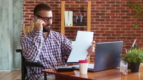 Disabled Freelancer in Wheelchair is Sitting at Table with Laptop Talking on Phone and Sorting