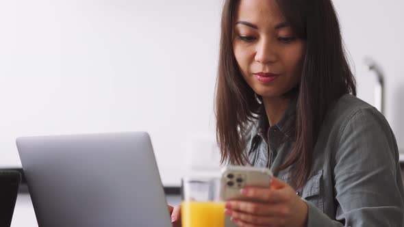 Pensive Asian woman looking at phone and starting to work by laptop