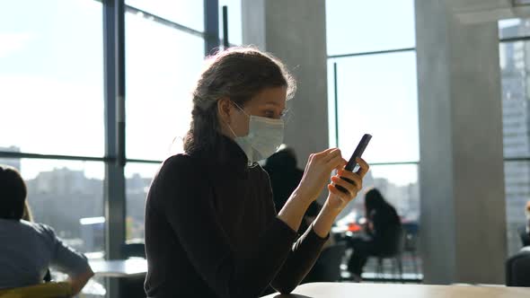 A Young Beautiful Girl in a Medical Mask Is Sitting with a Phone in a Shopping Center