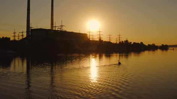 Silhouette of a factory near the river. Man in a boat roaming with oars at sunset. 