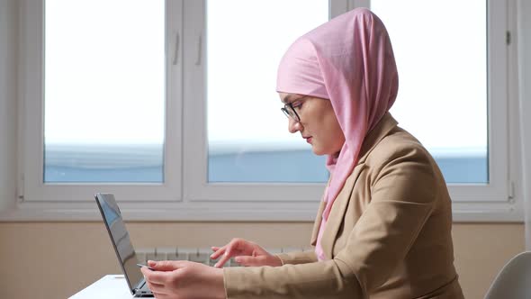 Side View of a Muslim Woman in a Pink Scarf Makes Online Purchases on a Laptop with a Card in Hands