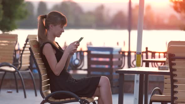 Young Woman Sitting at Street Cafe Table Talking on Her Cellphone Outdoors on Warm Summer Evening