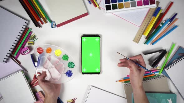 Man Holds Palette with Colours and Brush Over the Smartphone with Greenscreen alt
