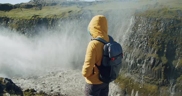 Man Hiker in Yellow Jacket and Backpack Standing at Cliff Edge Looking Down the Canyon with Mountain