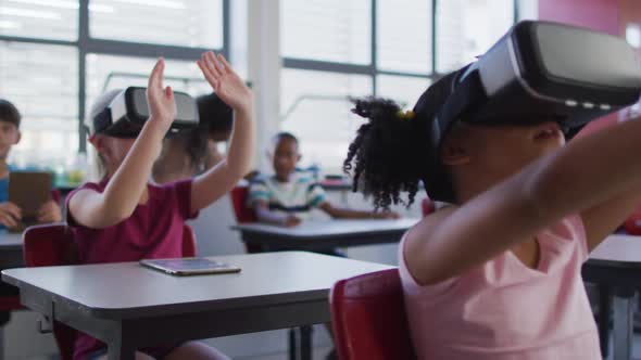 Diverse schoolchildren sitting in classroom wearing and using virtual reality glasses