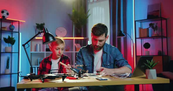 Father Soldering Important Details Under Lamp at Home in the Evening and His Interested 10-Aged Son