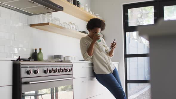 Happy african american woman drinking coffee and using smartphone in kitchen