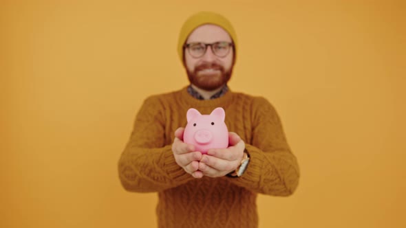 European Hipster Showing His Pink Piggy Bank to Camera Medium Studio Shot Yellow Background