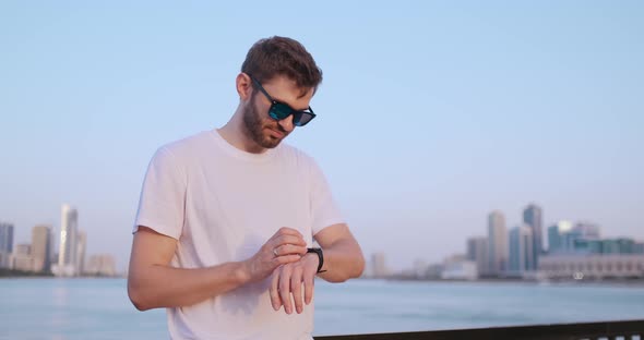 A Young Man Standing on the Waterfront in the Summer Uses the Smart Watch Screen