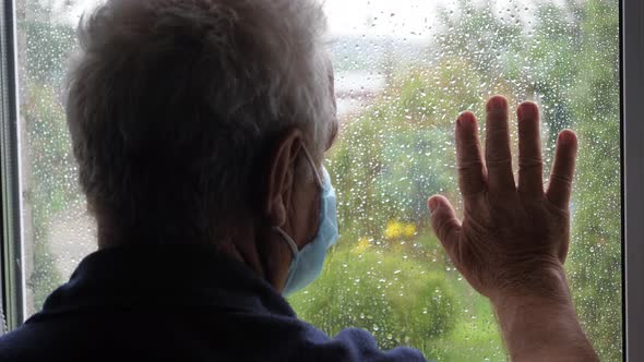 Old Man In Protective Medical Mask Looks Out Window And Puts His Hand On Glass